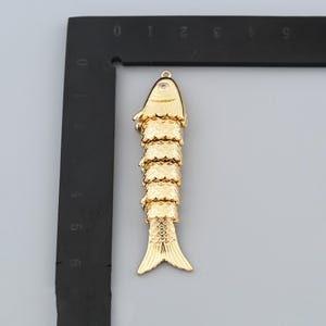 May include: A gold-colored fish pendant with a textured, scaled body and a small, clear gem near the top. The fish has a detailed fin and tail design, and a loop for hanging.