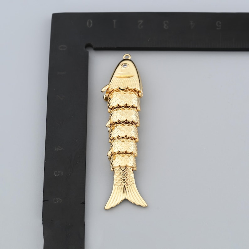 May include: A gold-colored fish pendant with a textured, scaled body and a small, clear gem near the top. The fish has a detailed fin and tail design, and a loop for hanging.