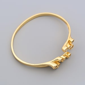 May include: A gold-colored open cuff bracelet. The bracelet has a simple, rounded band that widens at the ends, with a decorative, three-pronged design. The bracelet is made of a shiny, metallic material.