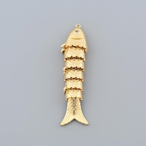 May include: A gold-colored fish pendant with a textured, segmented body and a loop at the top for hanging. The fish has detailed fins and scales, and a small eye detail. The pendant is likely made of metal.