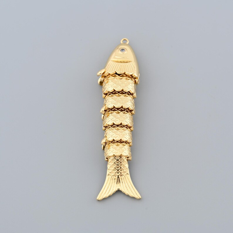 May include: A gold-colored fish pendant with a textured, segmented body and a loop at the top for hanging. The fish has detailed fins and scales, and a small eye detail. The pendant is likely made of metal.
