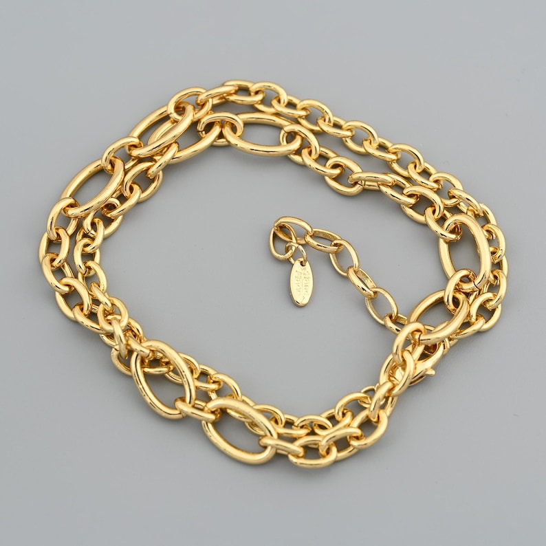 Puede incluir: Un collar dorado con un dise&ntilde;o de cadena de doble hebra. El collar presenta eslabones de forma ovalada y una peque&ntilde;a etiqueta cerca del cierre. La joya se exhibe sobre una superficie gris.