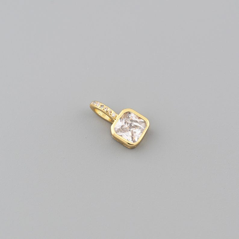 Peut inclure: Pendentif en or avec une pierre pr&eacute;cieuse carr&eacute;e et transparente. Le pendentif est dot&eacute; d'une b&eacute;li&egrave;re en or incrust&eacute;e de petites pierres transparentes. La pierre pr&eacute;cieuse est sertie dans une lunette en or. Fond gris.