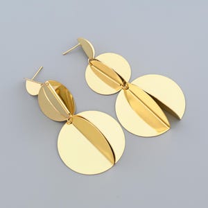 May include: A pair of gold-colored earrings with a post-back closure. Each earring features a combination of circular and leaf-shaped elements. The earrings have a polished, reflective surface and a modern, geometric design. The background is a solid, light gray.