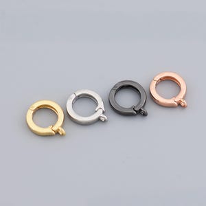 Könnte beinhalten: Vier runde Schmuckverschlüsse in Gold, Silber, Schwarz und Roségold. Jeder Verschluss hat ein kreisförmiges Design mit einer federbelasteten Öffnung und einer kleinen Schlaufe zur Befestigung an Schmuckkomponenten. Die Verschlüsse werden vor einem neutralen grauen Hintergrund gezeigt.