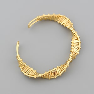May include: A gold-colored cuff bracelet with a woven design. The bracelet has an open design and is made of thin, rectangular pieces of metal woven together. The bracelet is on a gray background.