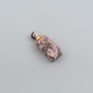 May include: A rectangular perfume bottle carved from pink and black patterned stone. The bottle features a gold-colored metal neck with two small loops and a matching stone stopper with a rounded top, showcasing natural mineral veining throughout.