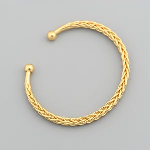 May include: A gold-colored cuff bracelet with a braided design. The open-ended bracelet features a rounded ball at each end. The bracelet is made of a shiny, metallic material and is set against a gray background.