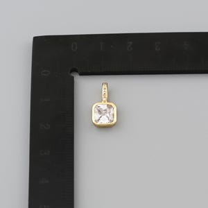 Peut inclure: Un pendentif dor&eacute; avec une pierre pr&eacute;cieuse carr&eacute;e et transparente. Le pendentif a une petite boucle en haut, orn&eacute;e de petites pierres transparentes. Le pendentif est &agrave; c&ocirc;t&eacute; d'une r&egrave;gle noire avec des marquages blancs.