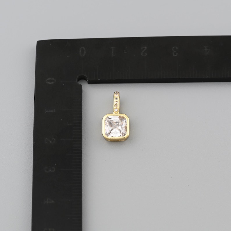 Peut inclure: Un pendentif dor&eacute; avec une pierre pr&eacute;cieuse carr&eacute;e et transparente. Le pendentif a une petite boucle en haut, orn&eacute;e de petites pierres transparentes. Le pendentif est &agrave; c&ocirc;t&eacute; d'une r&egrave;gle noire avec des marquages blancs.