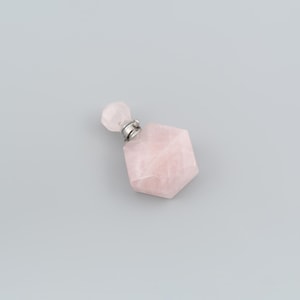 May include: A small, hexagonal-shaped perfume bottle carved from pale pink rose quartz. The bottle features a faceted surface and is topped with a matching rose quartz stopper, secured by a silver-toned metal collar at the neck of the container.