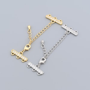May include: Two jewellery extenders, one gold-toned and one silver-toned, each with a bar of clear crystals. The extenders have a chain and a lobster clasp. The gold extender is on the left, and the silver extender is on the right.
