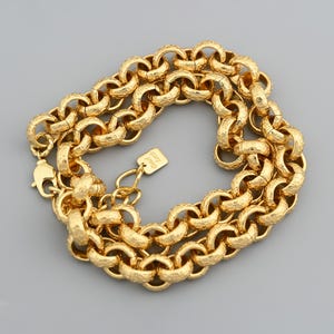 Puede incluir: Un collar de cadena dorado con eslabones redondeados texturizados. El collar tiene un cierre de mosquetón y una pequeña etiqueta que dice "Good Luck". La cadena se muestra sobre un fondo gris.