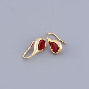 May include: A pair of gold-colored drop earrings with a teardrop-shaped, red gemstone. The earrings have a curved hook design for pierced ears. The gemstones are a deep, rich red color, set in a gold-tone metal.