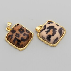May include: Two square pendants with a gold-colored, textured border. The pendants have a leopard print design in shades of brown and tan. Each pendant has a small gold loop at the top for attaching to a chain or cord.