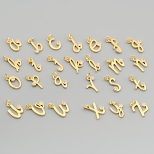 May include: A collection of gold-colored alphabet charms, each with a small, clear gemstone accent. The charms are in a cursive font and are displayed against a gray background. Each charm has a small loop for attaching to a chain or bracelet.
