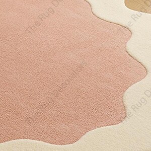 May include: Close-up of a soft, textured rug with a unique design. The rug features a large, irregular shape in a muted pink colour, bordered by a creamy off-white. The design has a wavy, organic edge.