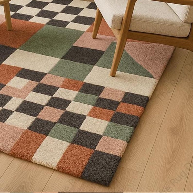 May include: A geometric patterned rug with a checkerboard and block design in shades of brown, green, pink, and cream. The rug is rectangular and has a soft, textured appearance. A wooden chair with a cream-colored cushion sits on the rug.