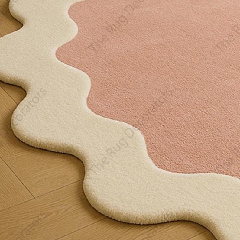 May include: A two-tone rug with a wavy edge, featuring a light pink central area and a cream-coloured border. The rug's design has a soft, cloud-like edge. The texture appears plush, suitable for a living space, with a modern aesthetic.