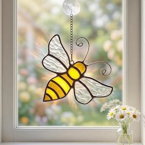 May include: A stained glass bee suncatcher with yellow and white wings, hanging in front of a window. The bee has a yellow body with black stripes. The wings are textured and translucent. A small vase of daisies sits on the windowsill.