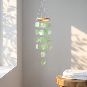 Peut inclure: Un mobile suspendu composé de multiples disques circulaires vert pâle translucides, fixés à un cadre en bois circulaire par de fines ficelles blanches. Les disques sont disposés en brins verticaux de longueurs variées, créant un effet en cascade.