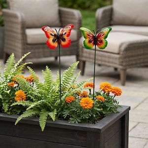 May include: Two metal butterfly garden stakes inserted into a rectangular wooden planter box. One butterfly features vibrant red and orange wings, while the other displays green and yellow wings. Both have black bodies and thin green metal stems.