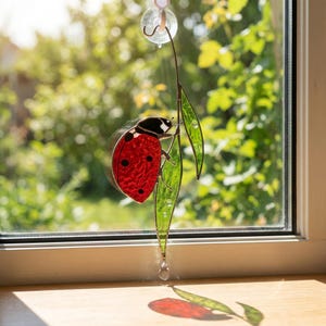 May include: A stained glass suncatcher featuring a red ladybug with black spots perched on a long, slender green leaf. The piece is suspended by a clear suction cup hook and includes a small clear crystal drop at the bottom of the leaf stem.