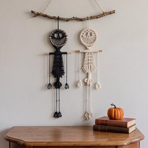 May include: Two macrame wall hangings shaped like skeletons, one in black and one in off-white, suspended from a natural wooden branch. Each figure features a circular head, a ribcage design, and long dangling limbs with knotted details.