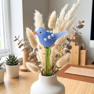 May include: A blue glass bird with white polka dots and an orange beak sits atop a green stem, placed in a white vase. The vase holds dried flowers and grasses, creating a decorative floral arrangement. The arrangement is on a wooden surface.