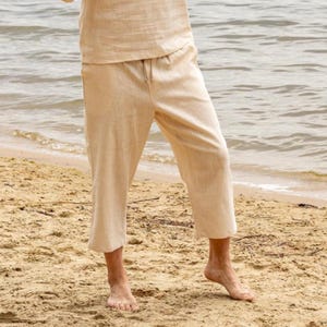May include: Beige linen trousers with a drawstring waist and cropped length. The trousers are part of a matching set, paired with a beige linen top. The outfit is worn on a sandy beach near the water.