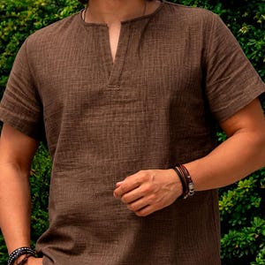 May include: A short-sleeved brown shirt with a textured fabric finish, featuring a round neckline with a vertical slit opening at the centre. The shirt is paired with white trousers and accessorised with layered bracelets on both wrists.