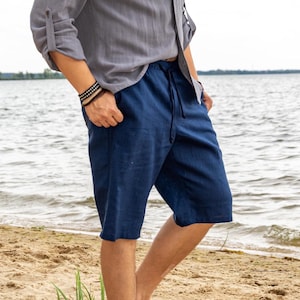 May include: Navy blue linen shorts with a drawstring waist. The shorts are knee-length and appear to be made of a lightweight fabric. The person is wearing a grey button-down shirt and a bracelet. The background shows a beach and water.