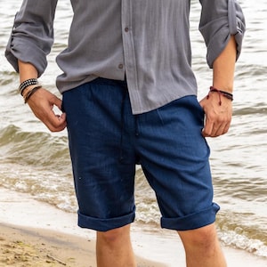 May include: Navy blue linen shorts with a drawstring waist and rolled cuffs. The shorts are paired with a grey button-down shirt with rolled-up sleeves. The outfit is casual and suitable for warm weather.