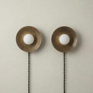 May include: Two wall sconces with a brass finish and a white globe light in the centre. Each sconce has a circular design and is connected to a black cord. The sconces are mounted on a light-coloured wall.