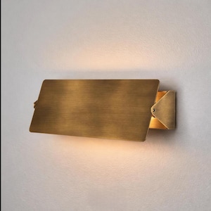 May include: A rectangular brass wall sconce with a hinged, angled shade. The sconce is mounted on a white wall, and the light is on, illuminating the area below. The fixture has a modern, minimalist design.