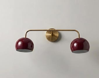 May include: A modern wall-mounted light fixture with two burgundy-colored, dome-shaped shades. The fixture has a brass-colored horizontal bar and a circular wall mount. The shades have a glossy finish and a silver-colored interior.