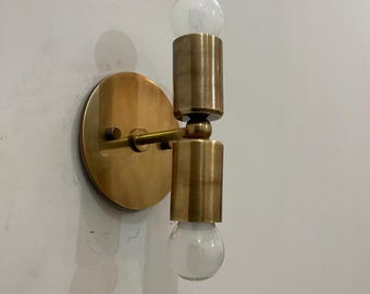 May include: A brass wall sconce with two clear glass light bulbs. The sconce has a circular backplate and cylindrical lamp holders. The fixture is mounted on a white wall, casting a shadow.