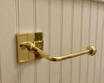 May include: A polished brass toilet paper holder featuring a square wall-mounted backplate with two visible screws and a horizontal arm that extends outward, ending in a rounded knob to secure a roll of paper in place.