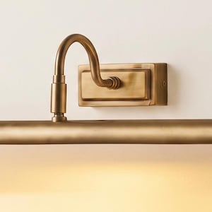May include: A wall-mounted picture light with a brushed brass finish. The fixture features a rectangular backplate, a curved arm, and a long, horizontal cylindrical lamp head that emits a warm, soft glow downward from the bottom edge.