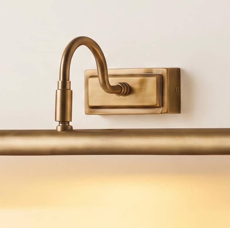 May include: A wall-mounted picture light with a brushed brass finish. The fixture features a rectangular backplate, a curved arm, and a long, horizontal cylindrical lamp head that emits a warm, soft glow downward from the bottom edge.