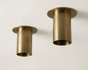 May include: Two cylindrical, brass-colored light fixtures with a circular base and a flared top. The fixtures are mounted on a white surface, and the design is minimalist and modern.