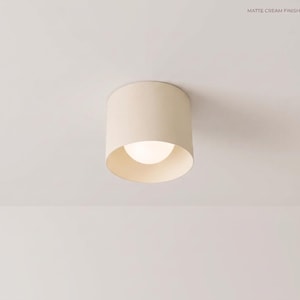 Può includere: Una lampada da soffitto cilindrica con finitura color crema opaco. Il dispositivo presenta un design a base larga e aperta che rivela una lampadina bianca sferica e luminosa centrata nell'alloggiamento. Il testo MATTE CREAM FINISH è visibile.