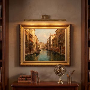 May include: A framed oil painting of a Venetian canal scene with buildings and boats, illuminated by a brass picture light. Below the painting sits a stack of books and a metal armillary sphere on a wooden surface between two tall bookshelves.