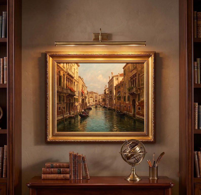 May include: A framed oil painting of a Venetian canal scene with buildings and boats, illuminated by a brass picture light. Below the painting sits a stack of books and a metal armillary sphere on a wooden surface between two tall bookshelves.