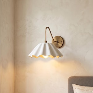 Op de afbeelding: Een witte keramische wandlamp met een geschulpte kap en een messingkleurige arm en achterplaat. De lamp is gemonteerd op een lichtgekleurde muur en de kap heeft een warme, uitnodigende gloed.
