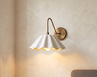 May include: A white ceramic wall sconce with a scalloped shade and a brass-colored arm and backplate. The light fixture is mounted on a light-colored wall, and the shade has a warm, inviting glow.