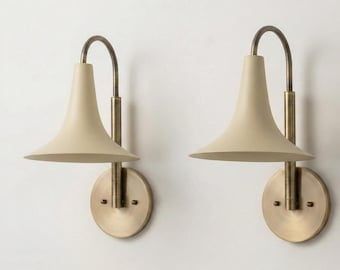 May include: Two wall sconces with a cream-colored shade and a brass finish. The sconces have a curved arm and a circular base. The shades are cone-shaped, and the overall design is modern and elegant.