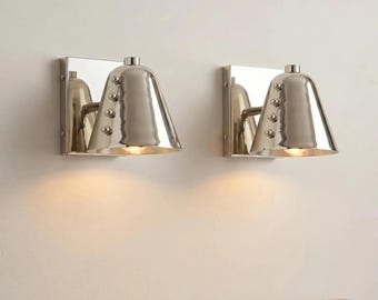 May include: A pair of polished silver-tone metal wall sconces, each featuring a square backplate and a conical shade. The shades are accented with a vertical row of four small spherical studs along the side and emit a warm downward light.