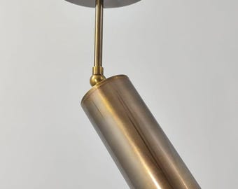 May include: A brass-toned, cylindrical pendant light fixture. The light has a circular ceiling mount with three screws, a straight rod, and a cylindrical light head. The light head is angled downward.