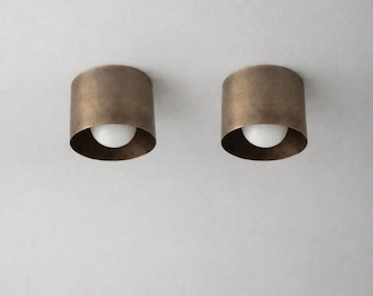 May include: Two cylindrical, bronze-colored light fixtures with a white, spherical light source. The fixtures are mounted on a white surface. The design is minimalist and modern, suitable for interior lighting.